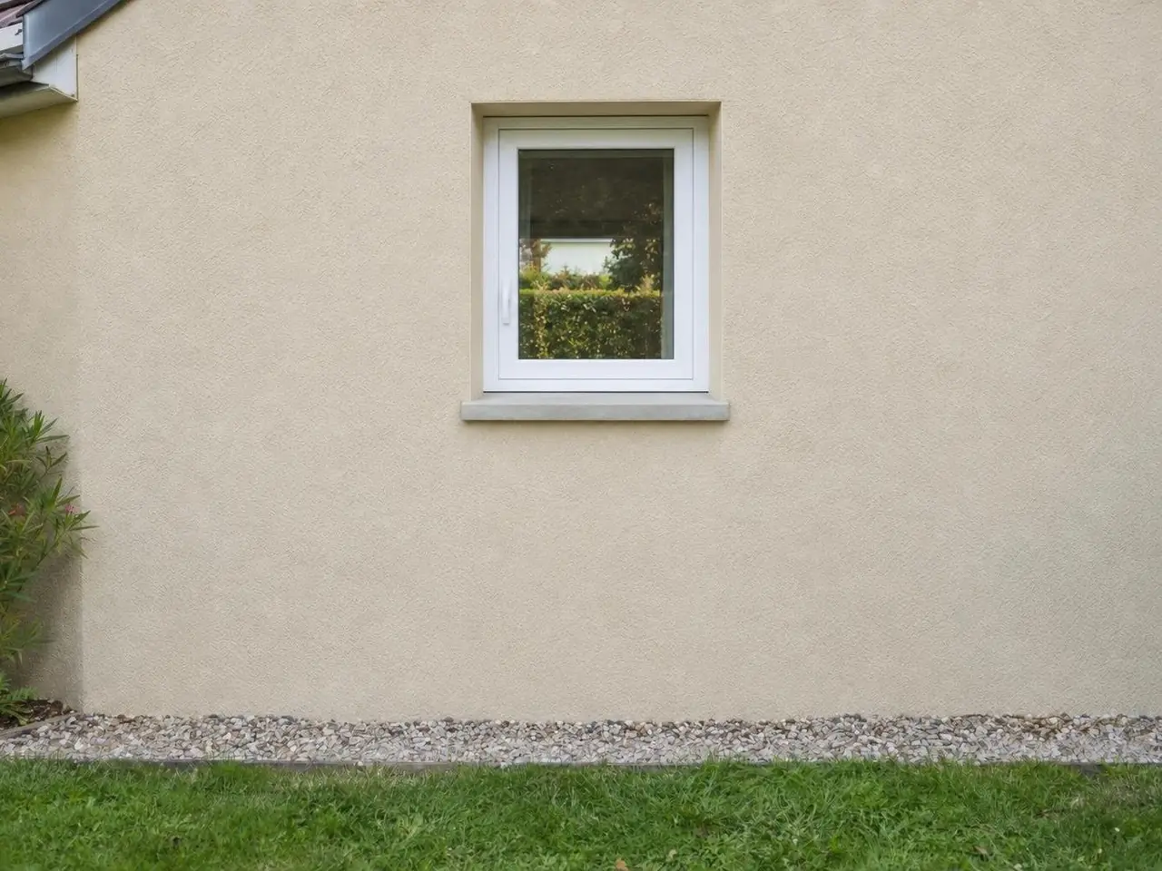 Façade en crépi beige percée d’une petite fenêtre blanche à un vantail, bande de gravier et pelouse au pied