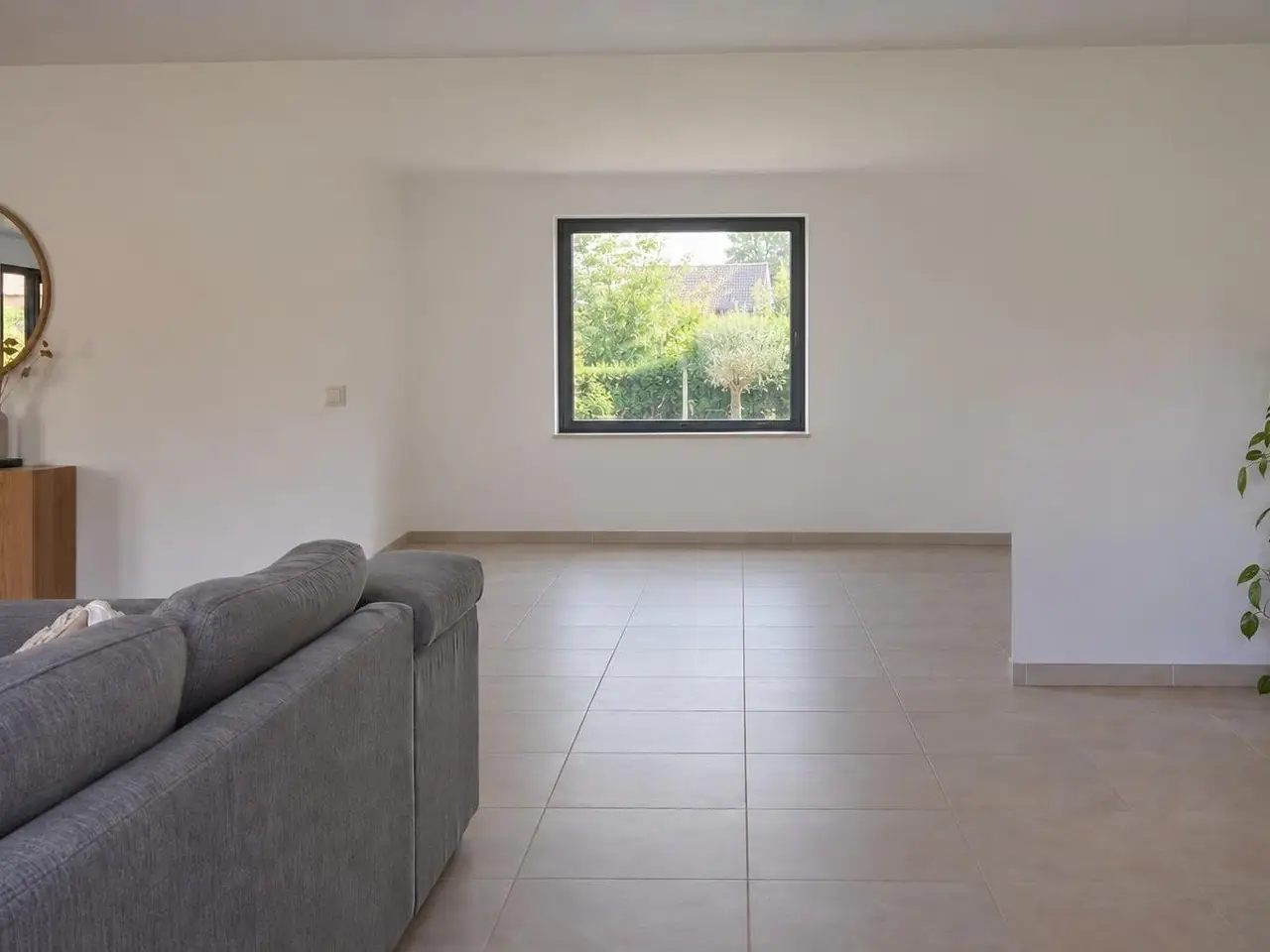 Séjour blanc au carrelage beige, canapé gris de dos, mur du fond plein percé d’une fenêtre carrée sur le jardin