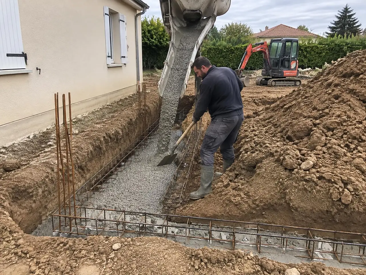 Toupie coulant le béton dans une fouille de fondation ferraillée le long d’un pavillon, maçon à la pelle