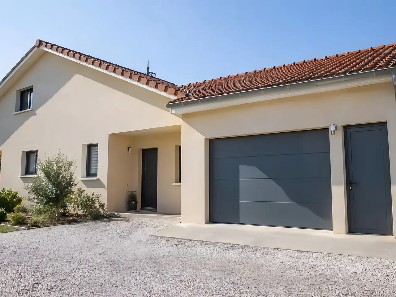 Pavillon enduit clair aux tuiles rouges, avec garage accolé à grande porte sectionnelle grise et porte de service