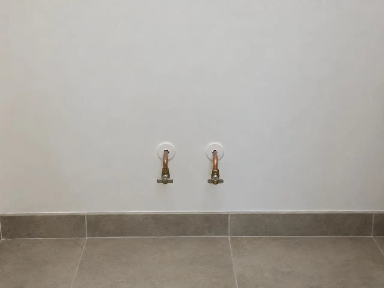 Mur blanc lisse de salle de bains, deux tuyaux en cuivre à robinets d’arrêt sortant de rosaces blanches, plinthe grise