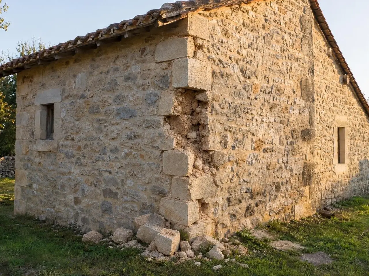 Angle d’une dépendance en pierre au toit de tuiles anciennes, pierres d’angle tombées dans l’herbe, blocage à nu