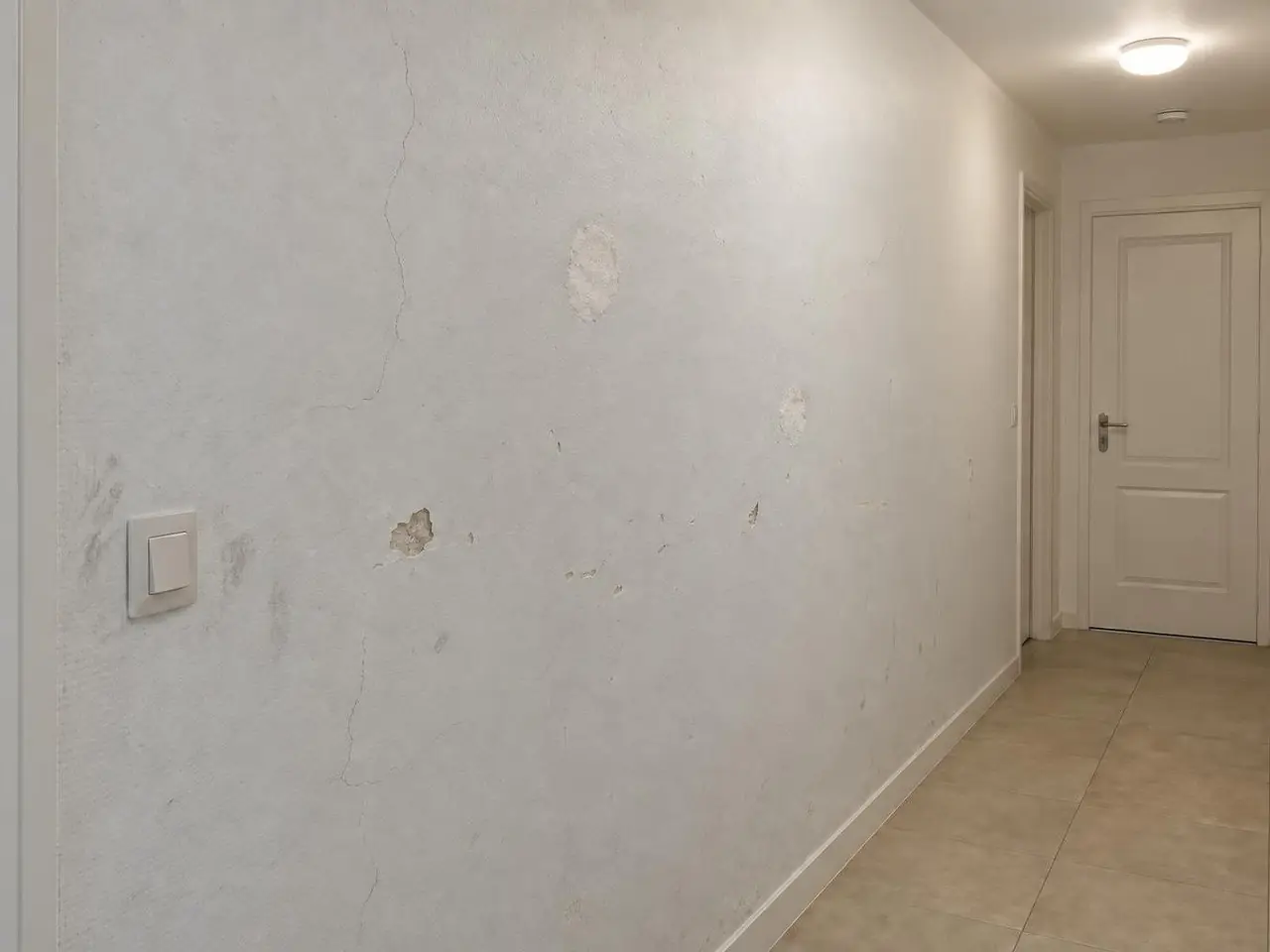 Mur de couloir peint en blanc grisé, fissures fines, éclats et rebouchages plus clairs autour de l’interrupteur