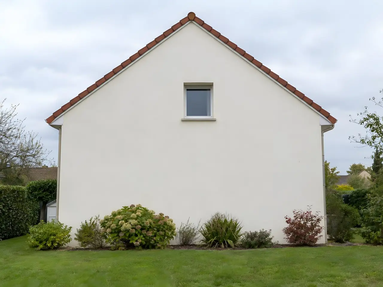Pignon d’un pavillon repeint d’un blanc cassé uniforme sous un toit de tuiles rouges, massif d’arbustes au pied