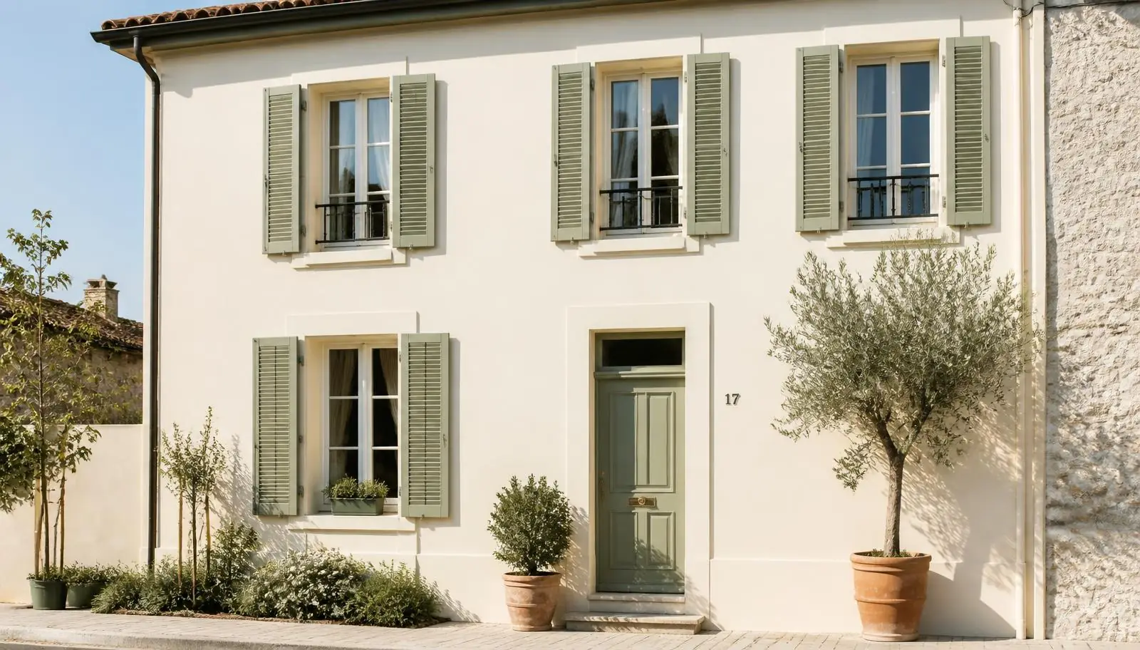 Façade de maison de village enduite crème, volets battants vert sauge, porte assortie et oliviers en pot