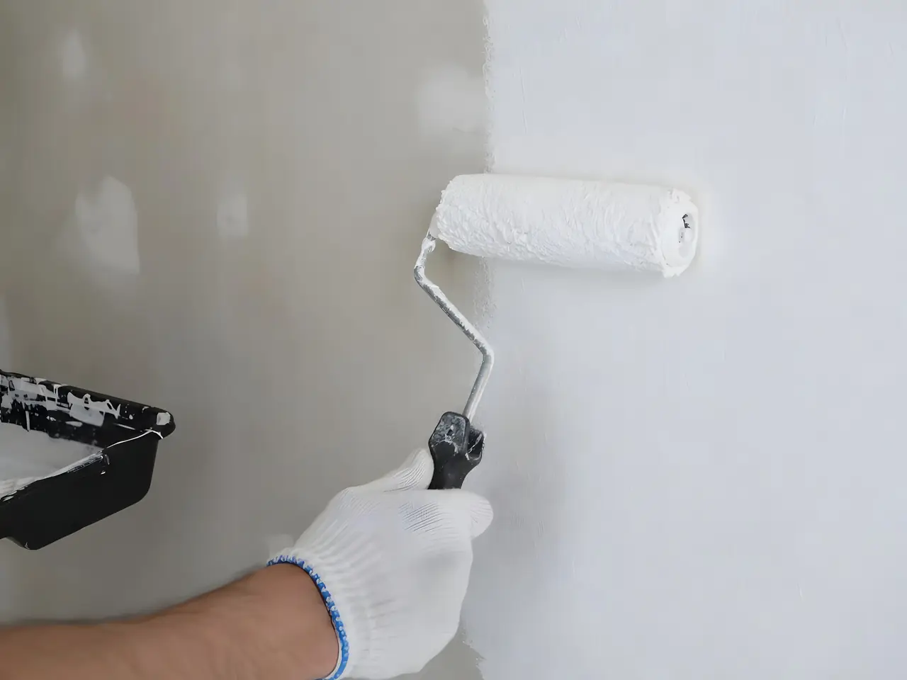 Main gantée qui passe un rouleau de peinture blanche sur un mur, la partie couverte nette à côté de l’enduit brut