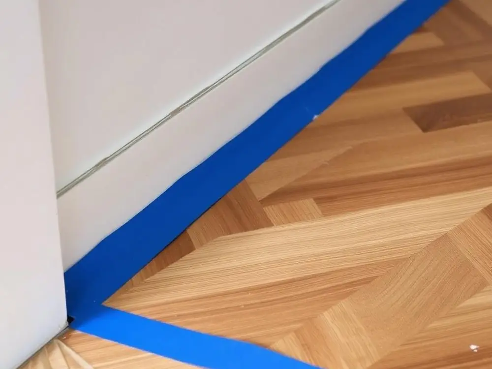 Adhésif de masquage bleu posé au ras d’une plinthe blanche, sur un parquet en bâtons rompus