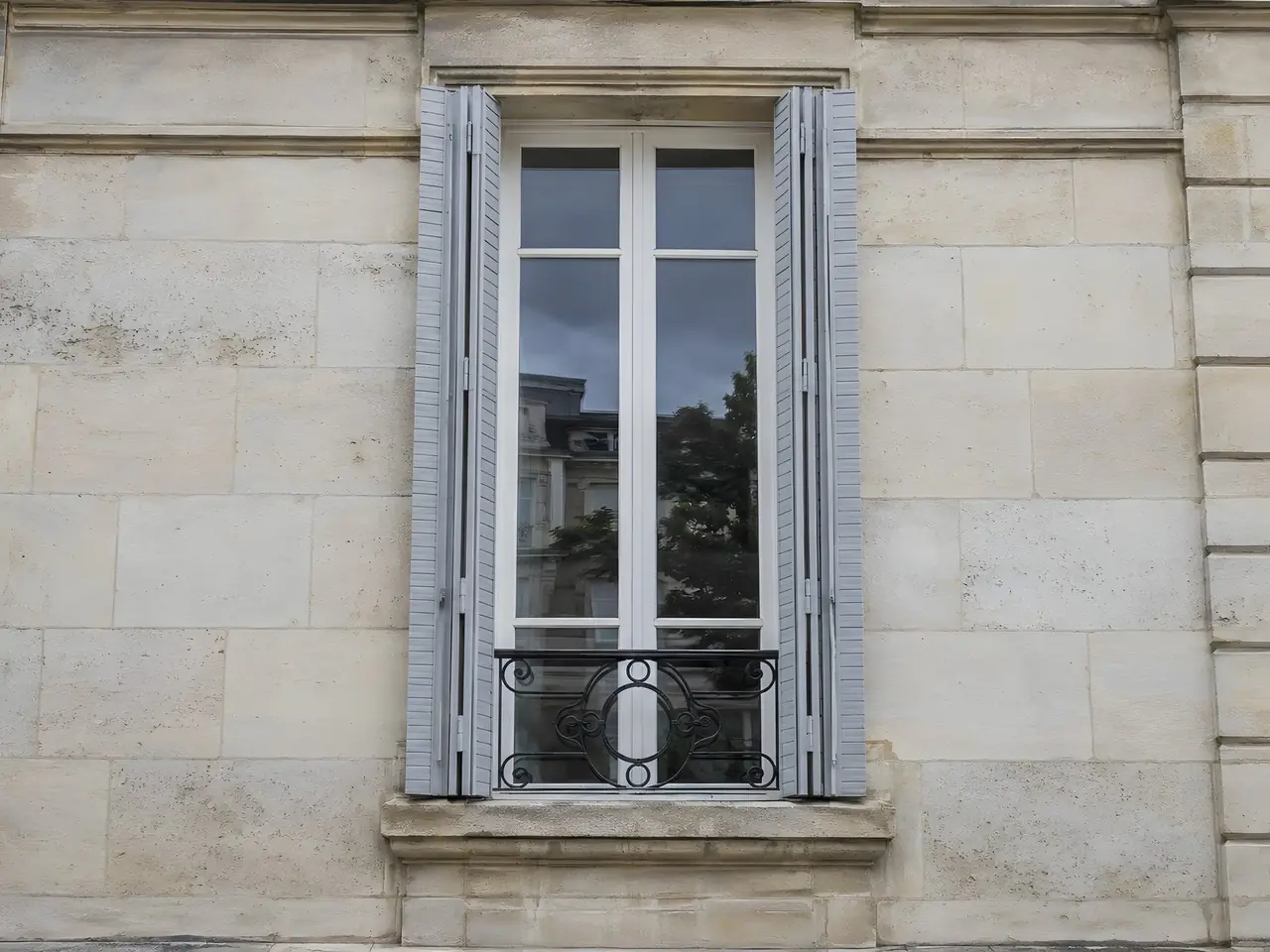 Persiennes métalliques repeintes en gris clair satiné, repliées dans le tableau d’une façade en pierre de taille