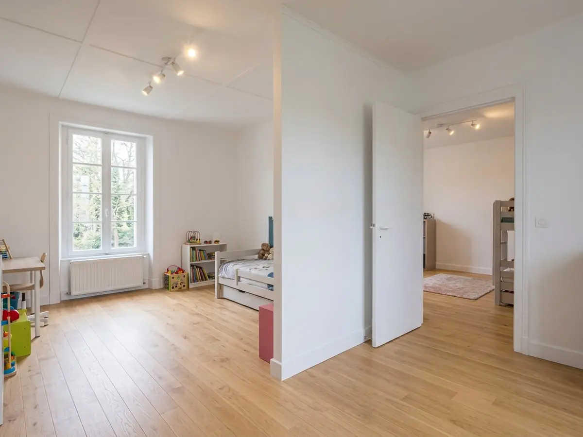 Cloison blanche qui partage une grande pièce en deux chambres d’enfant, porte ouverte de l’une à l’autre