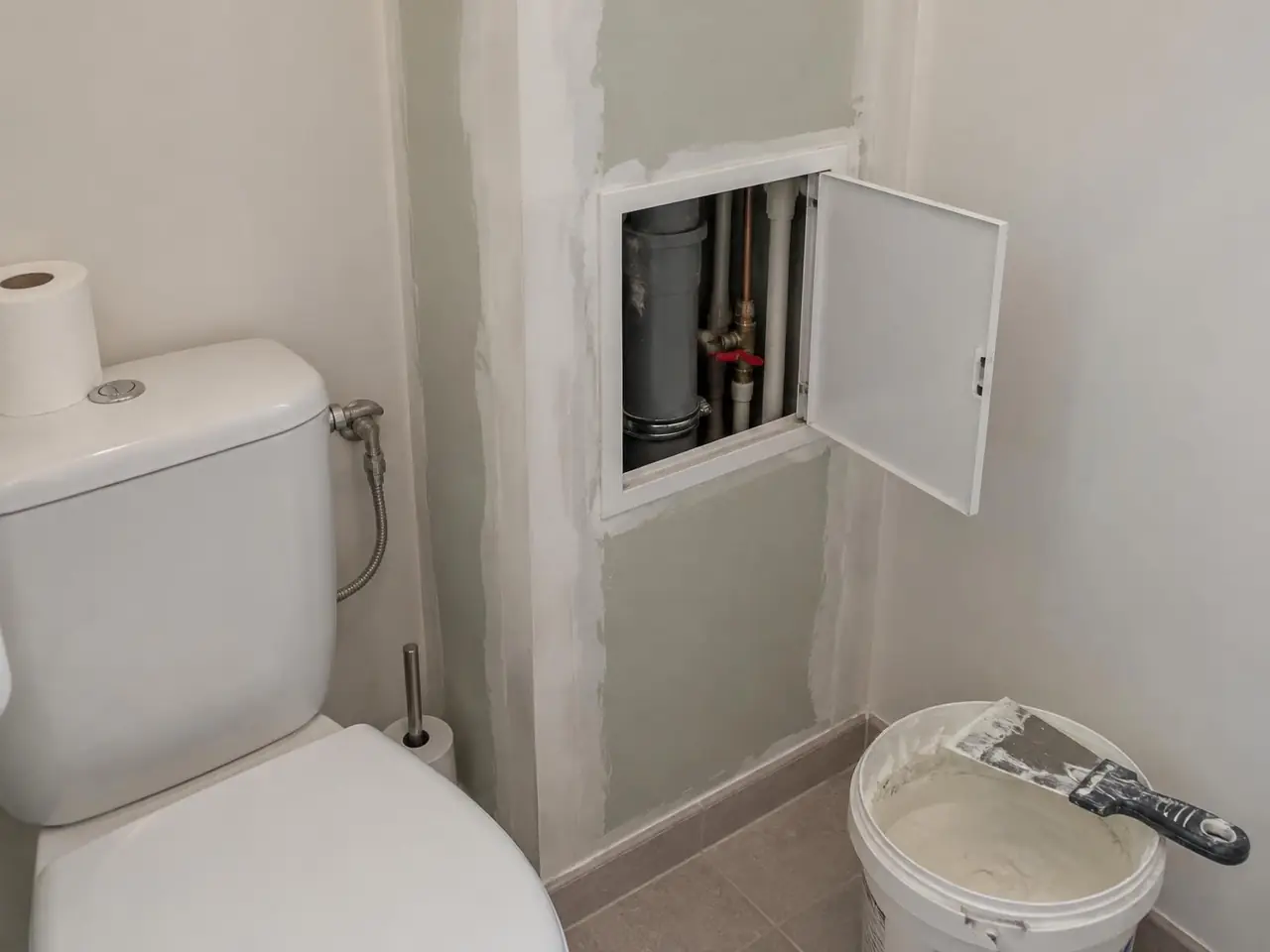 Coffrage en plaques dans des toilettes, trappe de visite ouverte sur la descente d’eau, les tuyaux et une vanne
