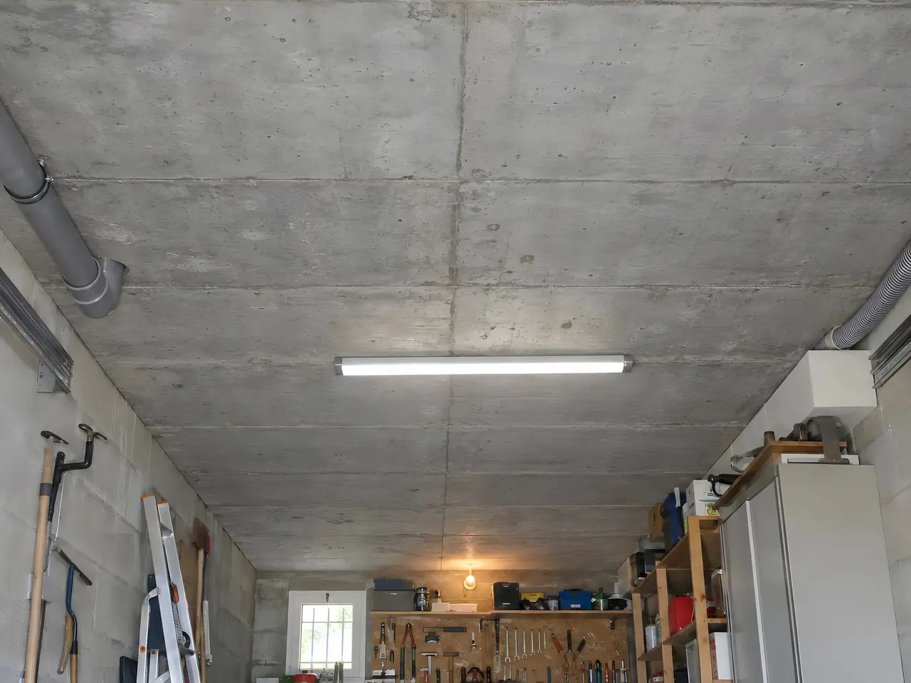 Plafond en béton brut d’un garage avec néon, gaines grises, échelle, outils au mur et établi au fond