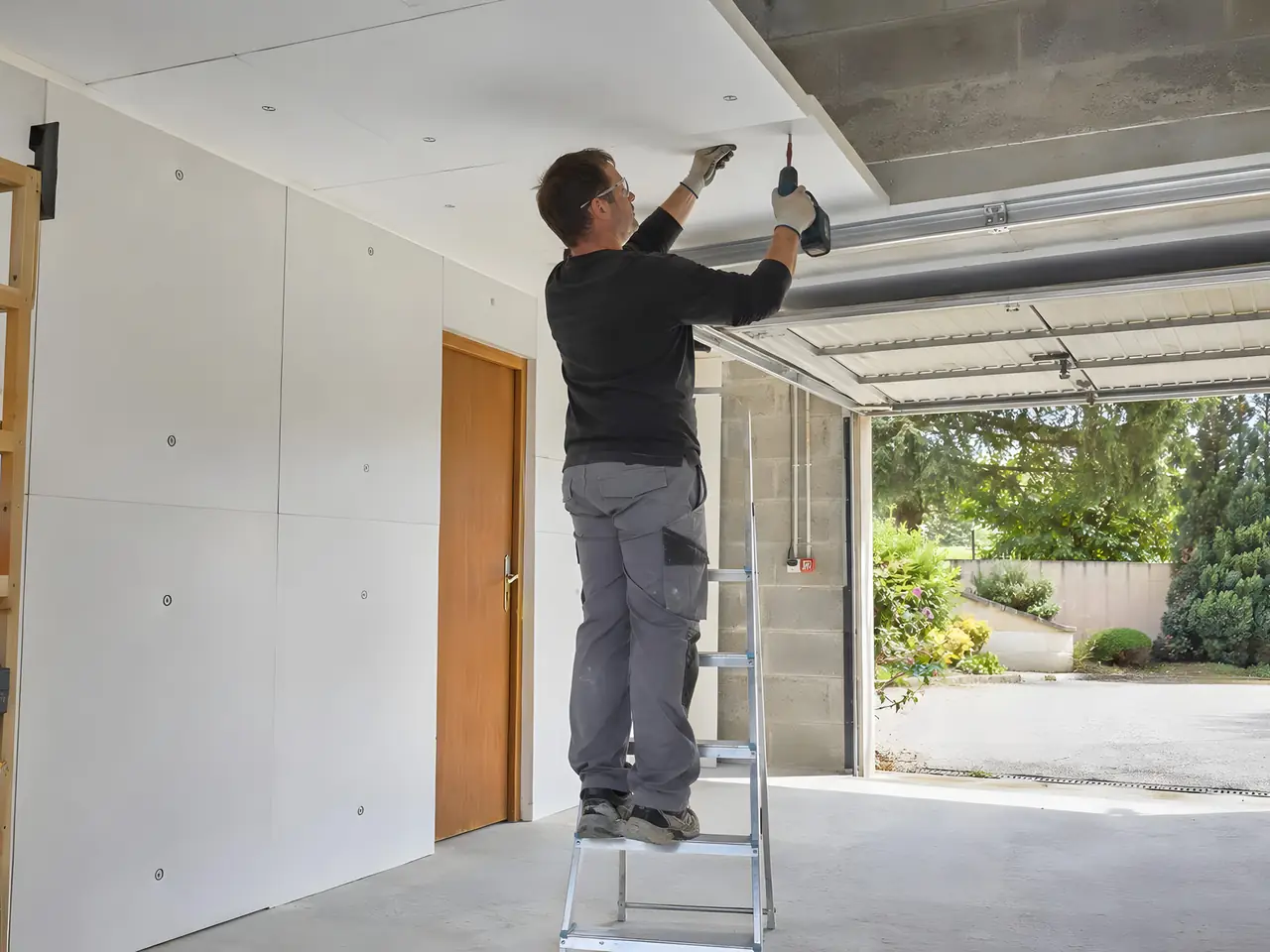 Artisan sur un escabeau qui visse des panneaux isolants blancs au plafond d’un garage, porte ouverte sur le jardin