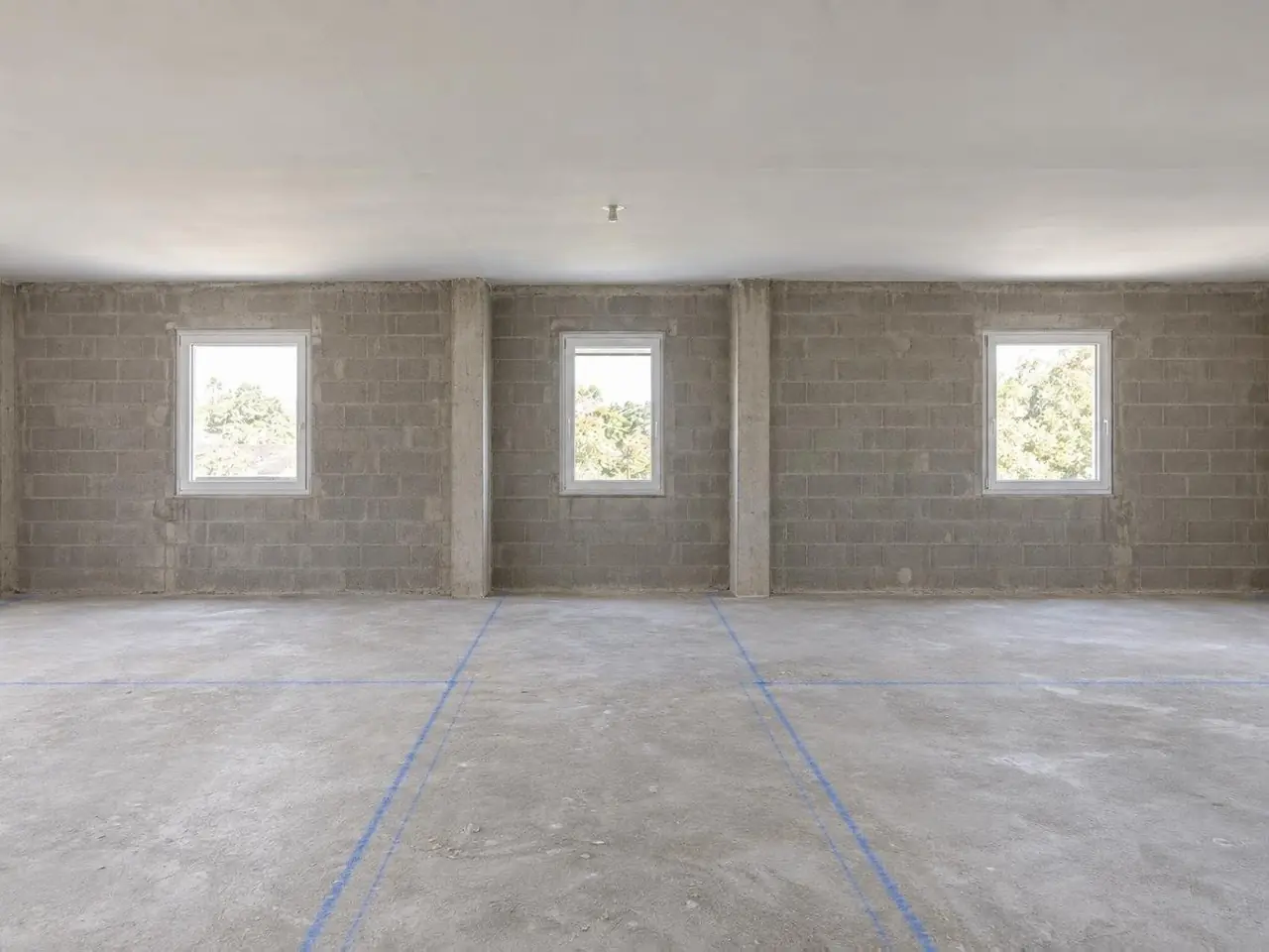 Étage de maison neuve vide aux murs de parpaings, trois fenêtres au fond, traits de cordeau bleus sur la chape