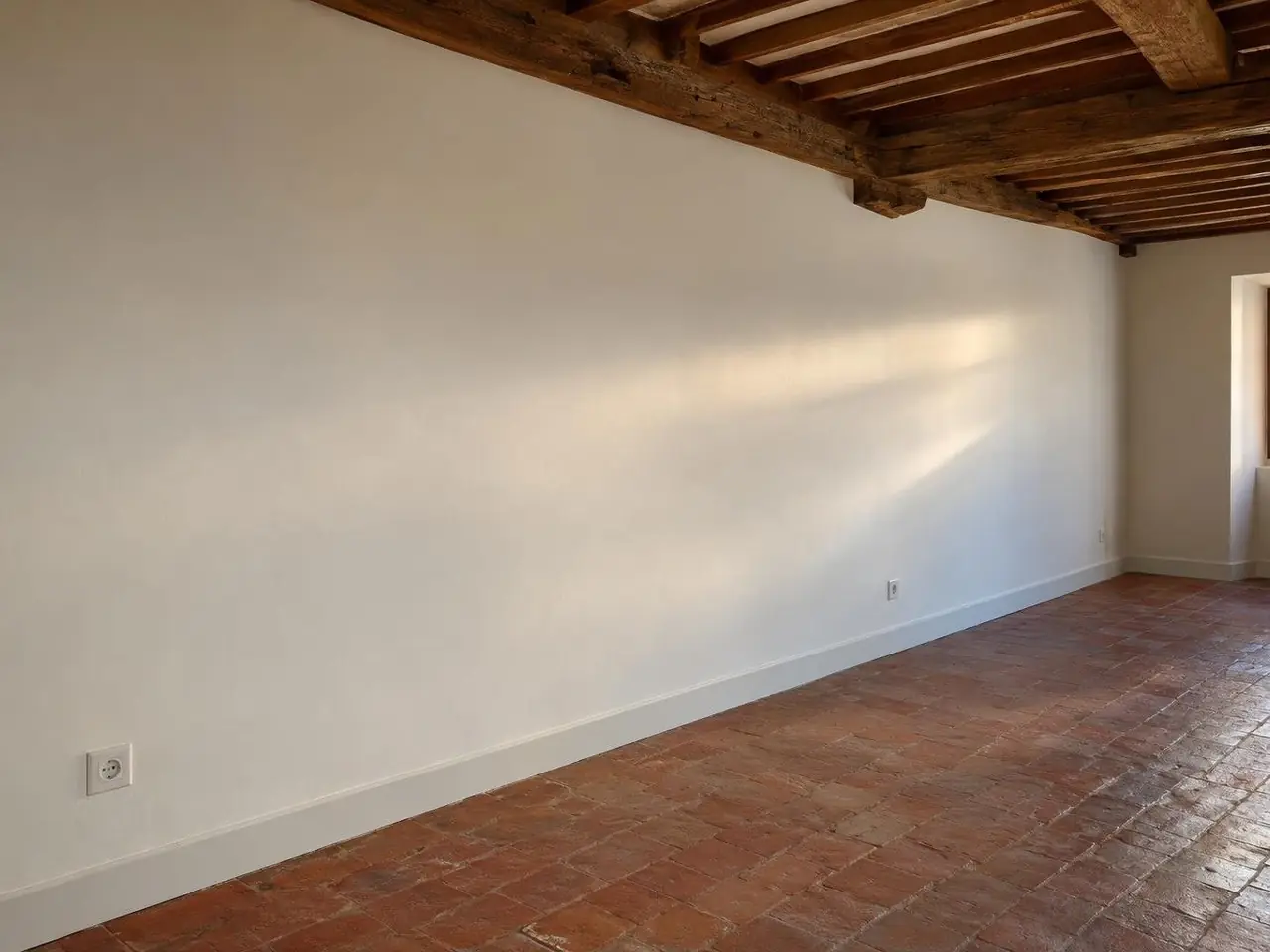 Long mur de séjour droit peint en blanc cassé, plinthe blanche rectiligne sous un plafond à poutres apparentes