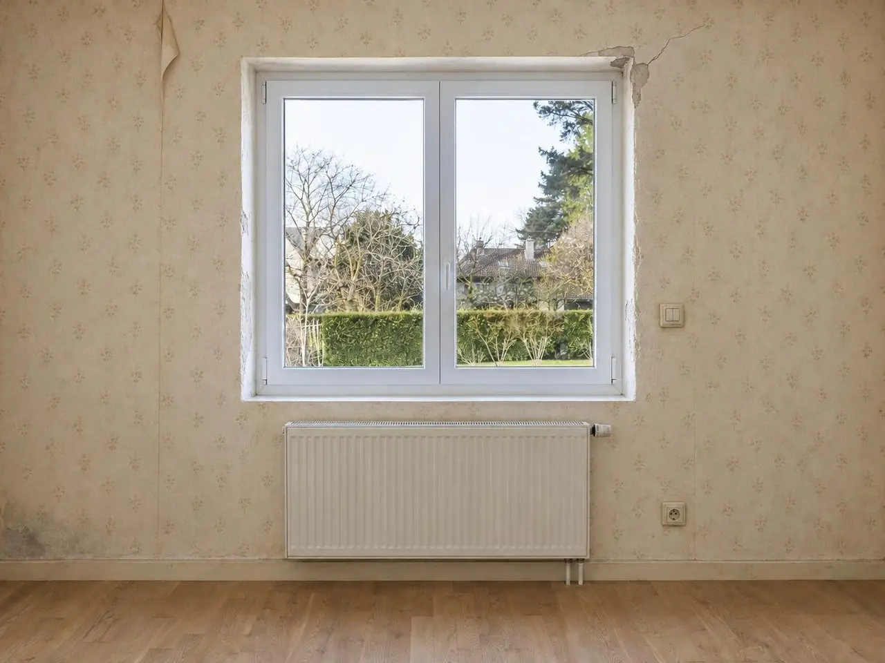 Mur de chambre au papier peint beige défraîchi, lé décollé en haut, fissure au coin de la fenêtre, radiateur