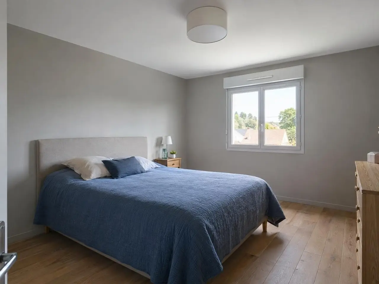 Chambre sous le grenier au plafond neuf d’un blanc uniforme, plafonnier rond, lit à couverture bleue