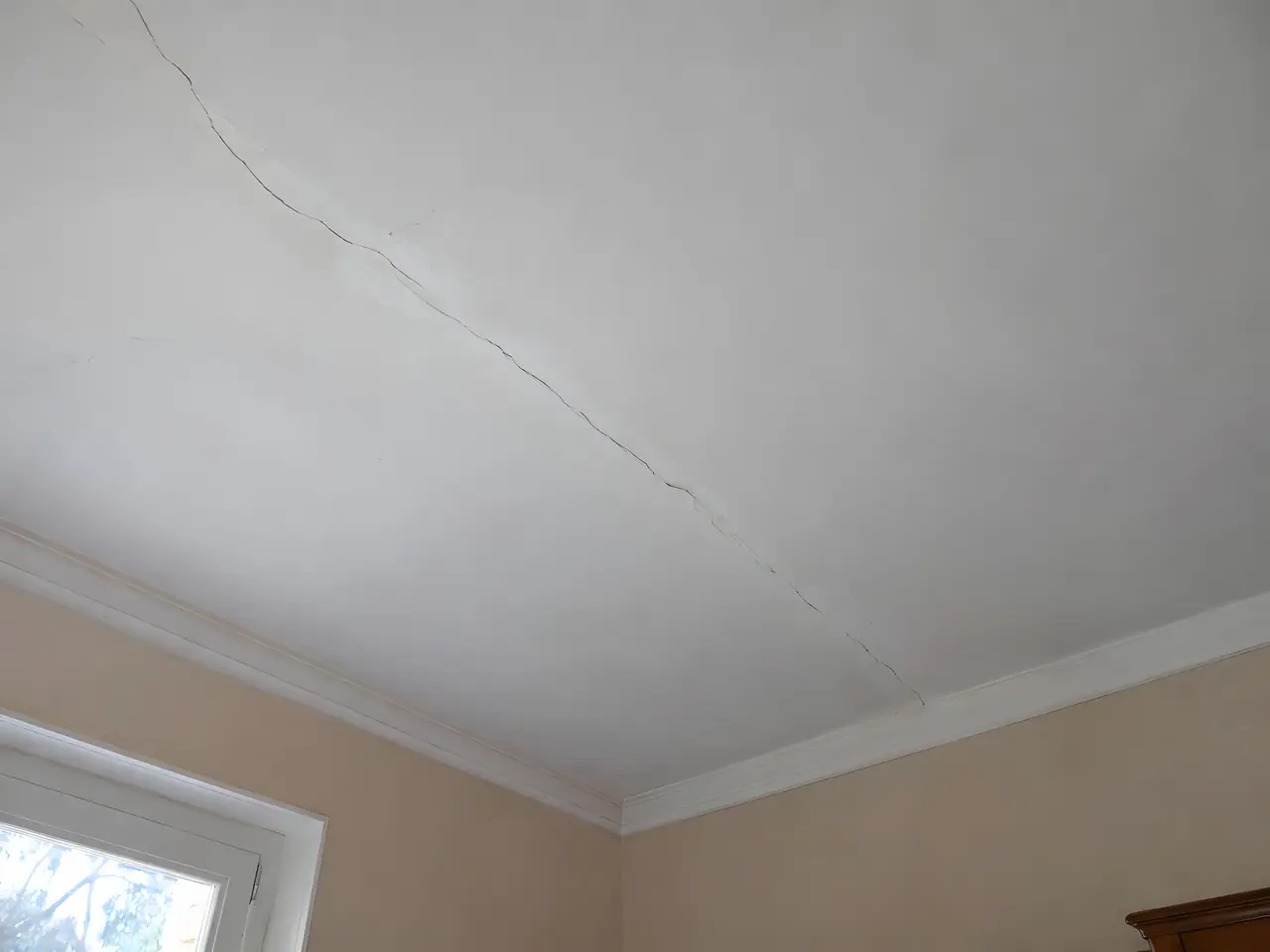 Plafond blanc ancien traversé d’une longue fissure en diagonale, au-dessus d’une corniche moulurée et de murs pêche