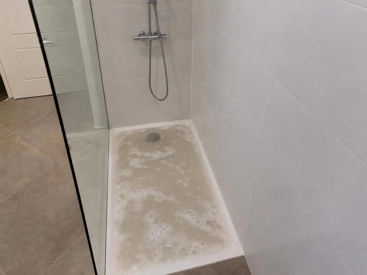 Receveur de douche blanc rempli d’eau grisâtre et savonneuse qui stagne, bonde à peine visible au fond