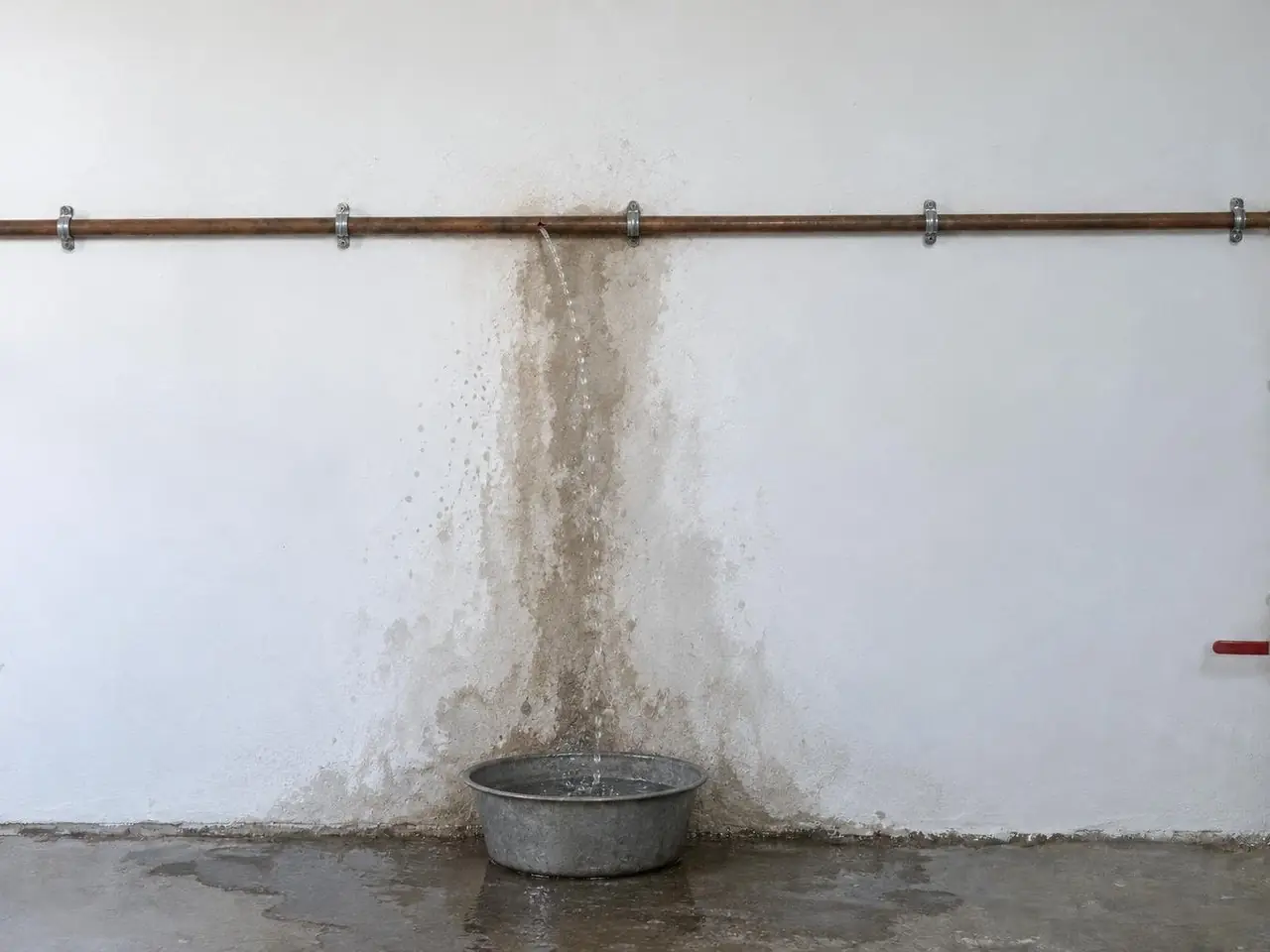Tuyau de cuivre fendu qui projette un filet d’eau sur un mur blanc taché d’humidité, bassine et flaque au sol