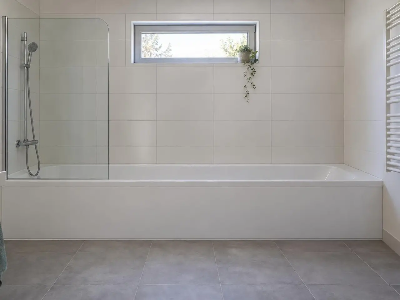 Salle de bains à carrelage blanc, longue baignoire à tablier blanc sous une fenêtre haute, pare-baignoire vitré