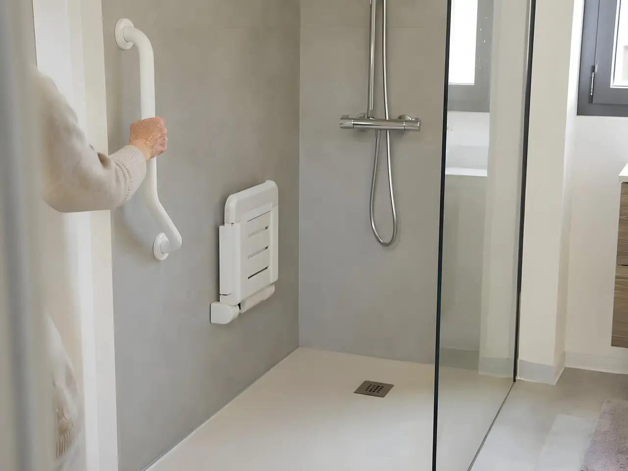 Douche de plain-pied avec barre d’appui blanche, siège rabattable et paroi vitrée, une main âgée tenant la barre