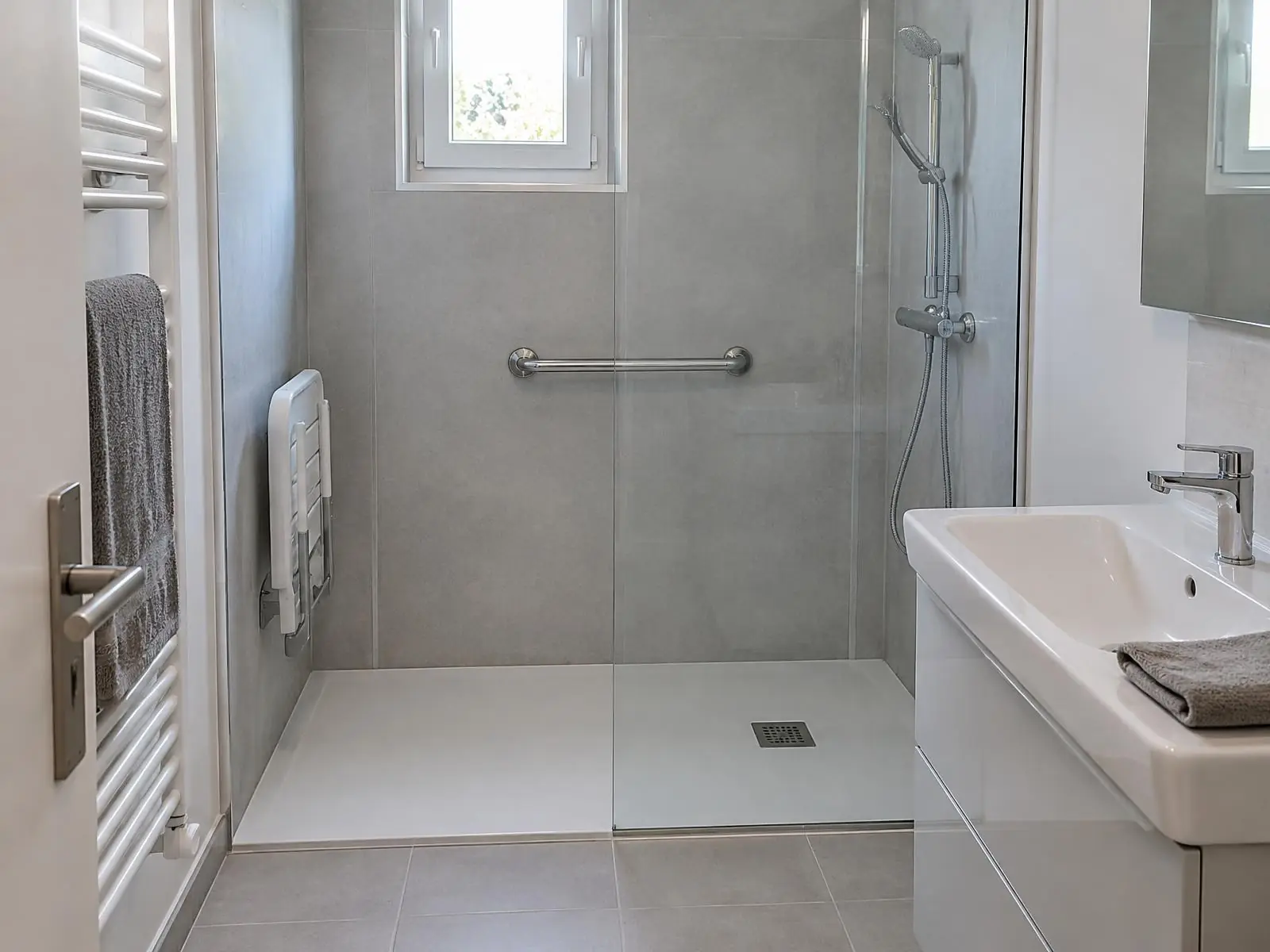 Douche extra-plate derrière une paroi vitrée, barre d'appui et siège relevable au mur gris, lavabo à côté