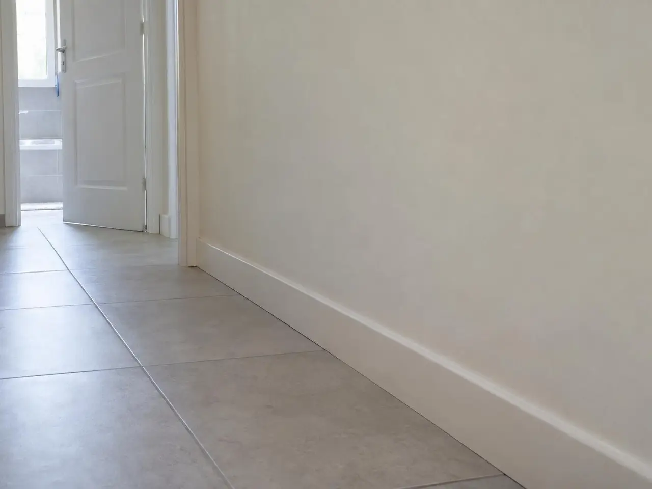 Bas de mur de couloir peint en blanc cassé, propre et uni, plinthe blanche droite sur un carrelage gris clair