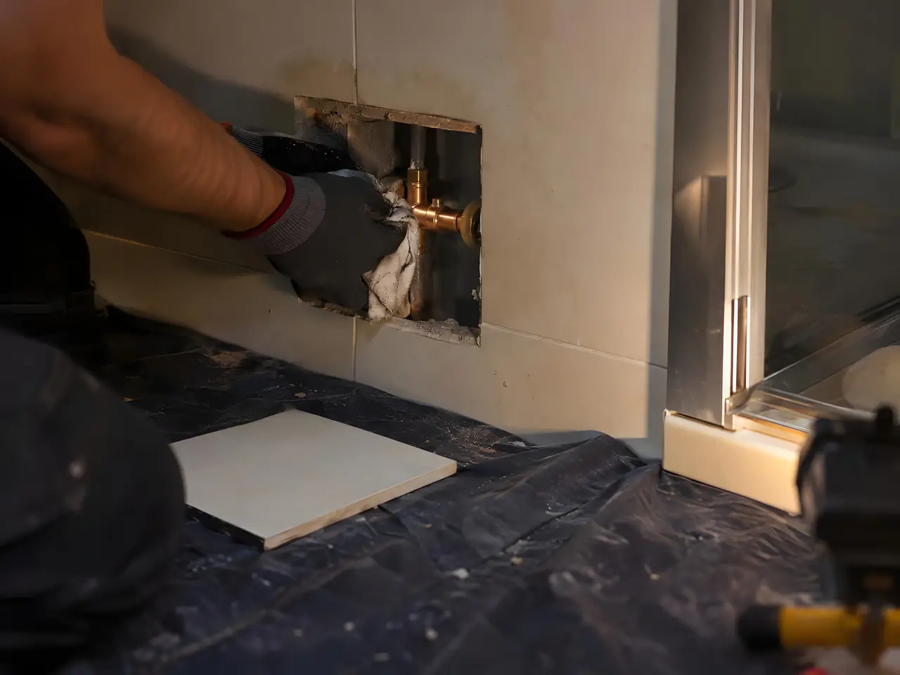 Main gantée qui essuie un raccord en laiton par une trappe ouverte dans la faïence, au pied d’une douche
