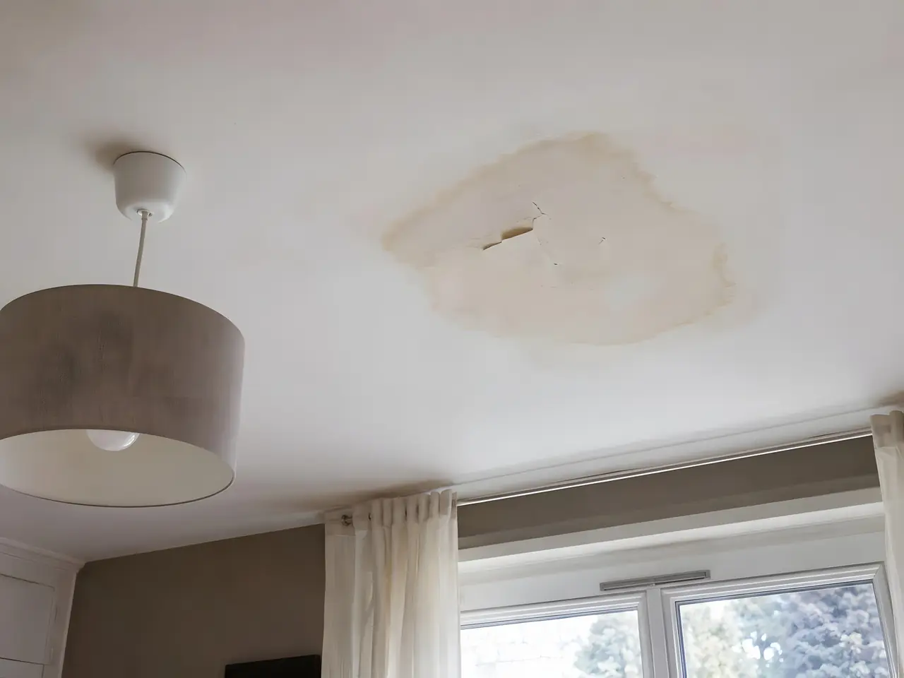 Plafond blanc marqué d’une grande auréole jaunâtre de dégât des eaux, près d’une suspension en tissu gris