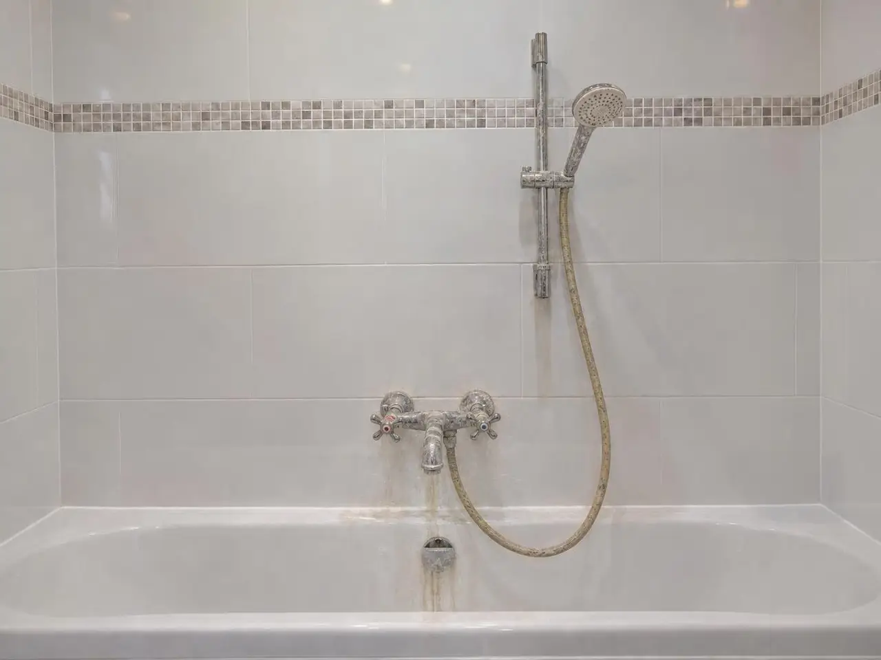 Vieux mélangeur de baignoire à deux têtes couvert de calcaire, flexible jauni, coulure sur l’émail