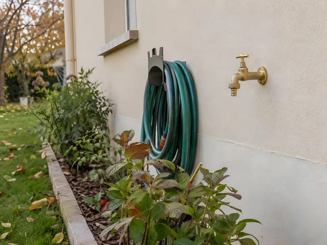 Robinet extérieur en laiton neuf sur une façade enduite beige, tuyau d’arrosage vert enroulé sur son support