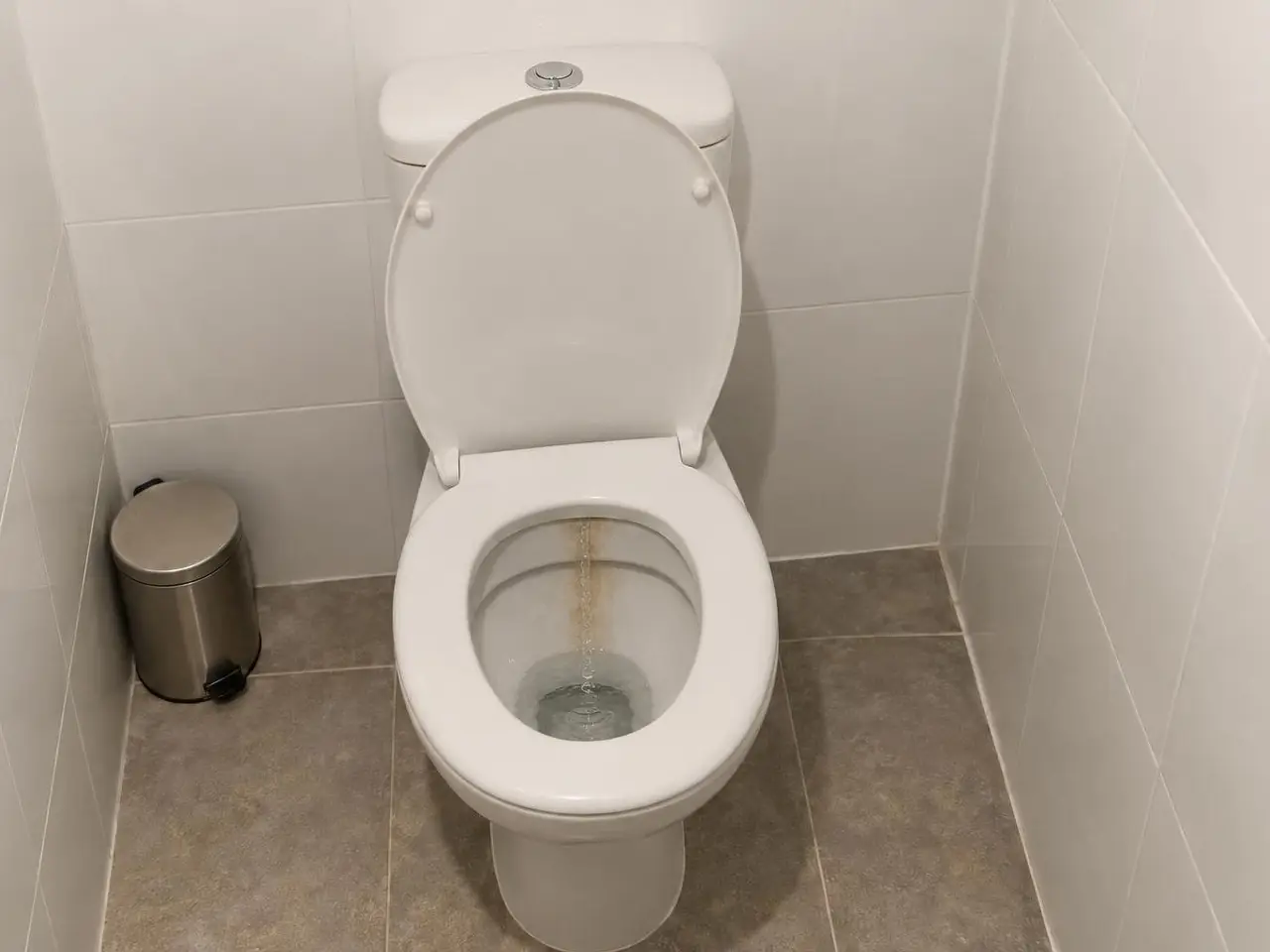 Cuvette de WC blanche abattant relevé, traînée orangée de calcaire le long de la paroi arrière, eau qui coule